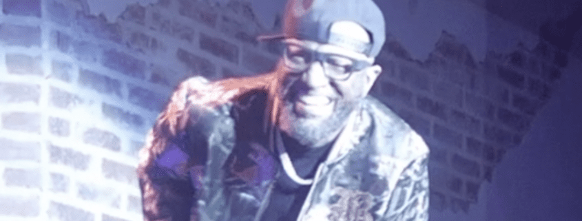 Man smiling with cap, brick wall background.